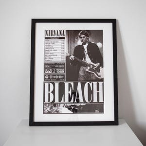 Peut inclure: Affiche encadrée en noir et blanc de l'album "Bleach" de Nirvana. L'image montre un membre du groupe jouant de la guitare, avec le titre de l'album et la liste des titres. L'affiche est dans un cadre noir et repose sur une surface blanche.