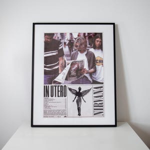 Könnte beinhalten: Ein gerahmtes Poster mit einem Foto der Band Nirvana, auf dem drei Mitglieder zu sehen sind, von denen eines eine Zeitung liest. Darunter stehen die Texte IN UTERO und NIRVANA, eine Titelliste und eine Grafik einer geflügelten Figur mit dunklen Federn.