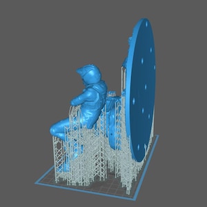 May include: A light blue 3D-printed model of a seated astronaut figure with a large circular component. The astronaut is wearing a helmet and a spacesuit. The model is supported by a lattice structure.