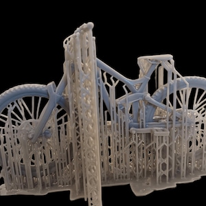 May include: A light blue mountain bike model, 3D printed with support structures. The bike features detailed wheels, frame, and components, showcasing the intricate design. The model is set against a black background.