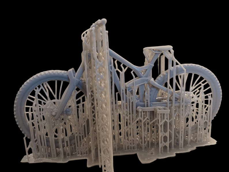 May include: A light blue mountain bike model, 3D printed with support structures. The bike features detailed wheels, frame, and components, showcasing the intricate design. The model is set against a black background.