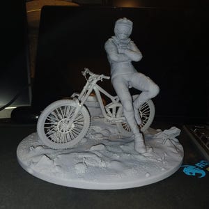 May include: A gray figurine of a person in a racing suit, leaning against a mountain bike. The figure is on a circular base with a textured surface, suggesting a terrain. The bike has detailed wheels.