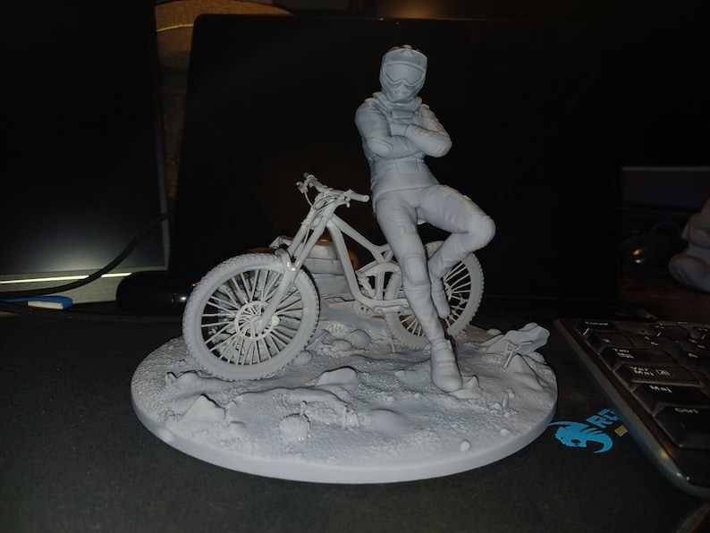 May include: A gray figurine of a person in a racing suit, leaning against a mountain bike. The figure is on a circular base with a textured surface, suggesting a terrain. The bike has detailed wheels.
