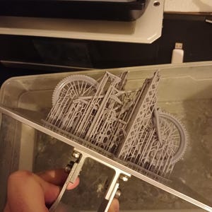 May include: A gray 3D-printed model of a bicycle, held by a metal support. The intricate design includes wheels, frame, and other details. The model is partially submerged in a liquid, likely during the 3D printing process.