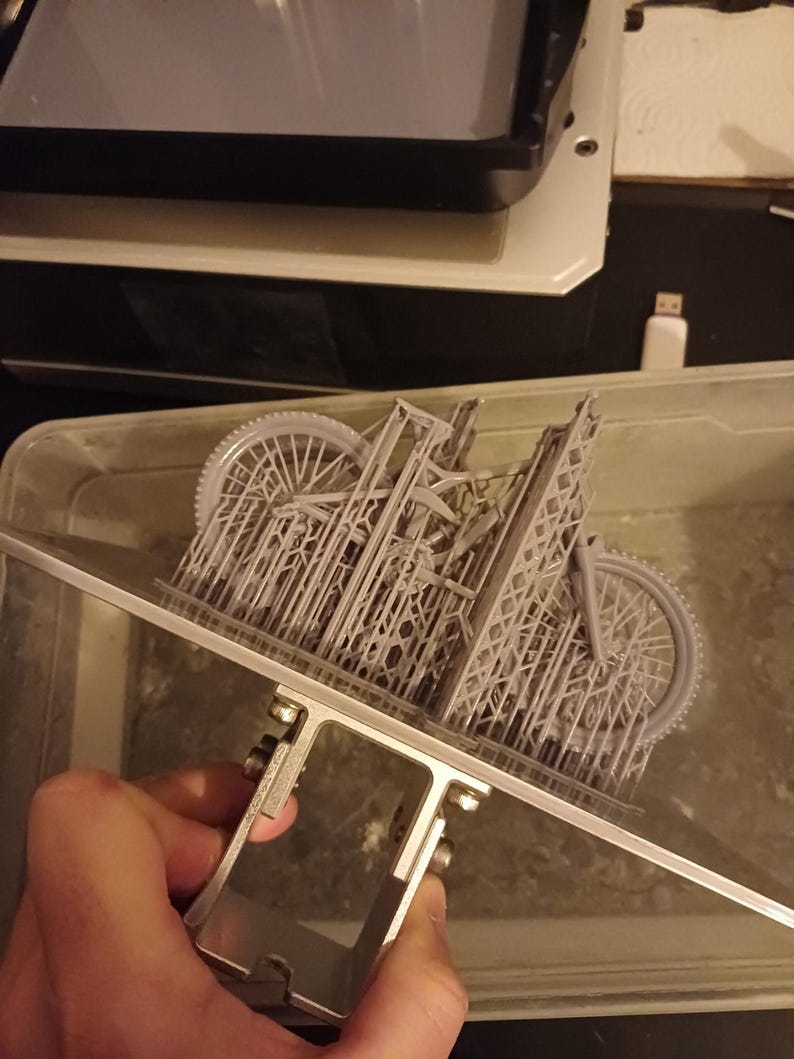 May include: A gray 3D-printed model of a bicycle, held by a metal support. The intricate design includes wheels, frame, and other details. The model is partially submerged in a liquid, likely during the 3D printing process.