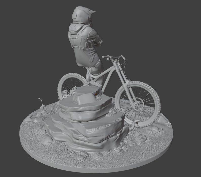 May include: A gray sculpture of a mountain biker on a bicycle, posed on a rocky terrain. The biker is wearing a helmet and protective gear. The bicycle has large tires and suspension. The sculpture is on a circular base.