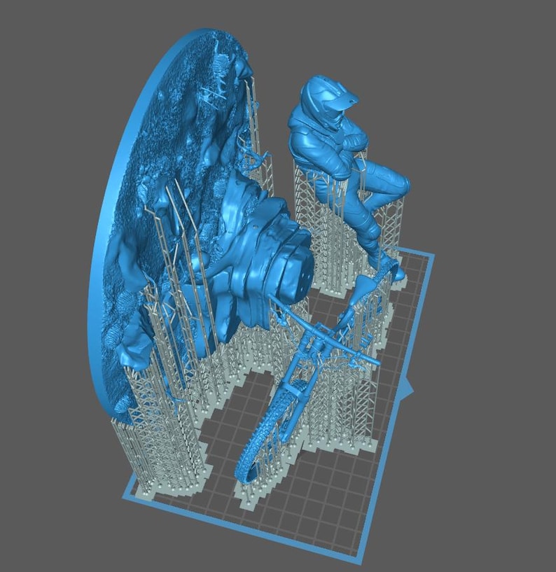 May include: A blue 3D-printed model featuring a motorcycle, a rider in a helmet, and a detailed landscape element. The model is supported by a grid-like structure, suggesting it's in the printing process. The overall theme is likely related to motorsports or model-making.