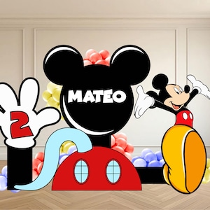 Op de afbeelding: Een grote feestuitsparing met Mickey Mouse-thema, voorzien van een zwart silhouet van een hoofd met de naam MATEO in witte letters, een rode basis met twee blauwe ovale ramen en een witte gehandschoende hand die een rood cijfer 2 vasthoudt.