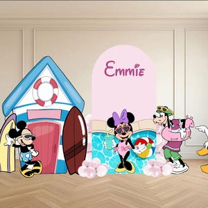May include: A decorative party backdrop set featuring a blue and white beach house, a pink arched panel with the name Emmie, and cartoon characters including Mickey, Minnie, Pluto, Goofy, and Daisy Duck in summer attire with beach accessories.