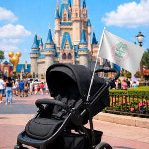 Puede incluir: Un cochecito negro con una bandera blanca con el logo "Enchanted Pop Designs" en rosa y verde. El fondo muestra un castillo y una multitud, lo que sugiere un parque tem&aacute;tico.