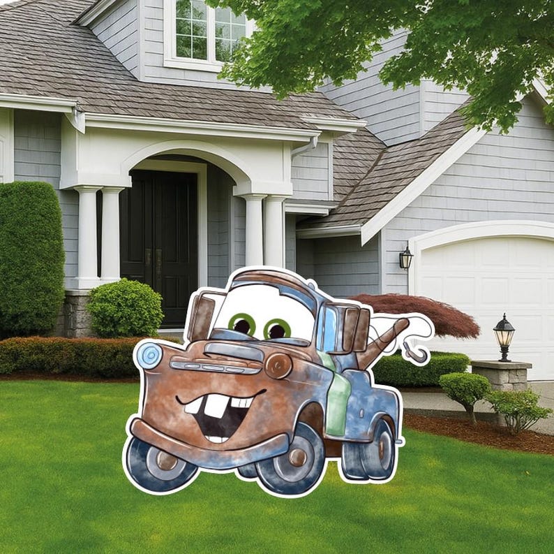 May include: A decorative yard sign featuring a cartoon-style rusty brown tow truck with large green eyes, a wide smiling mouth with two prominent front teeth, and a towing hook on the back, displayed on a green lawn in front of a house.
