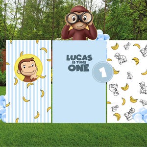 May include: A party backdrop featuring a cartoon monkey with binoculars. The backdrop includes three panels: one with the monkey and bananas, one with the text "LUCAS IS TURNS ONE", and one with monkey and banana patterns. Blue and white balloons are at the base.