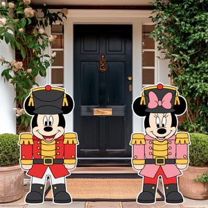 May include: Two decorative nutcracker-style cutouts featuring cartoon mice characters. The left figure wears a red uniform with gold accents and a black hat. The right figure wears a pink uniform with gold accents, a pink bow, and a black hat.