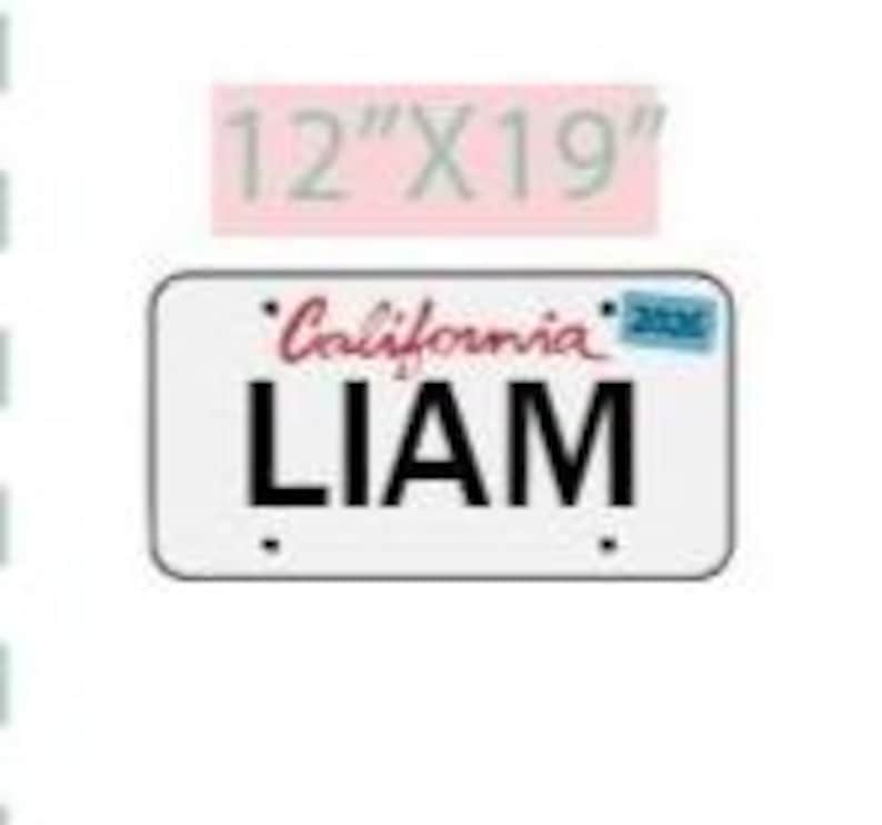 May include: A white license plate with the name "LIAM" in bold black letters. The word "California" is above the name in red script. The plate measures 12" x 19".