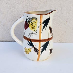 May include: Cream-colored ceramic pitcher with a curved handle and spout. The pitcher is decorated with brown, yellow, and dark green leaf and fruit designs. Two brown bands encircle the pitcher.