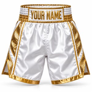 May include: White satin boxing shorts featuring a wide, gold-trimmed waistband with the text "YOUR NAME" in gold lettering. The shorts include metallic gold snakeskin-textured side panels and gold trim along the leg openings.
