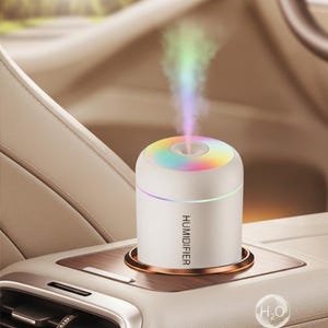 Op de afbeelding: Een witte luchtbevochtiger met een regenboogkleurige bovenkant en het woord "HUMIDIFIER" op de zijkant. Het apparaat geeft kleurrijke damp af en staat in de console van een auto. De basis is koperkleurig.