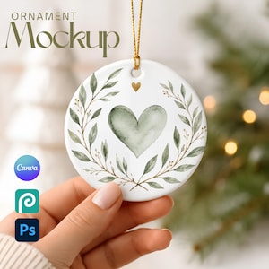 May include: A round white ceramic ornament featuring a watercolour sage green heart centred within a delicate botanical wreath of green leaves. A small gold heart sits above the main heart. The ornament hangs from a thin gold string.