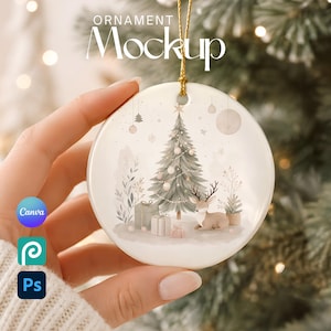 May include: A round white ceramic Christmas ornament featuring a printed illustration of a decorated pine tree, wrapped gift boxes, and a small deer. The ornament hangs from a gold string and displays the text ORNAMENT MOCKUP at the top.