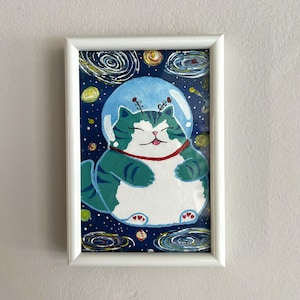 Può includere: Un dipinto incorniciato di un gatto tigrato rotondo, verde e bianco, che indossa un casco spaziale trasparente. Il gatto ha un collare rosso e fluttua su uno sfondo spaziale blu scuro pieno di galassie vorticose, stelle gialle e piccoli pianeti colorati.