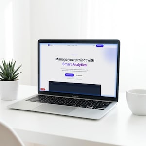 Puede incluir: Un portátil plateado que muestra una interfaz de sitio web con el texto "Manage your project with Smart Analytics". Una pequeña suculenta en una maceta blanca y una taza blanca están sobre el escritorio blanco. El portátil está abierto.