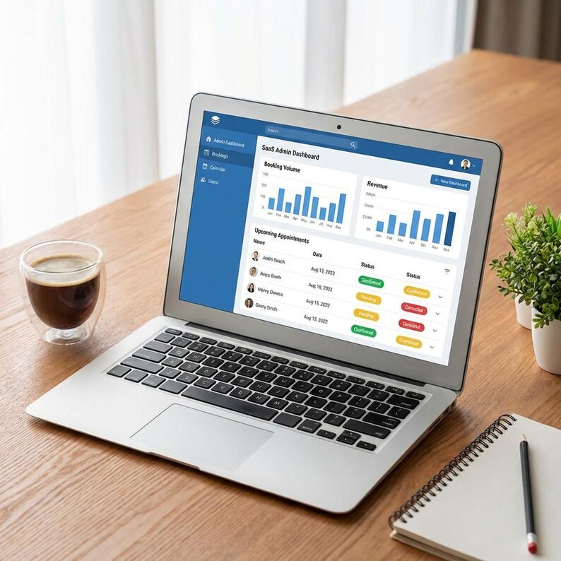 May include: A silver laptop displaying a SaaS Admin Dashboard with booking volume and revenue charts. A cup of coffee, a small potted plant, and a notepad with a pencil are on a wooden table. The laptop screen shows upcoming appointments.