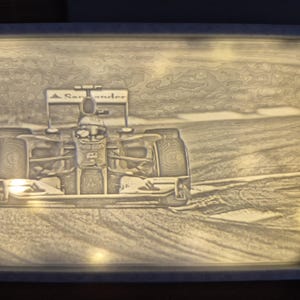 Op de afbeelding: Een verlicht, ingelijst kunstwerk met een Formule 1-raceauto. De grijswaardenafbeelding toont de auto in beweging, met het Santander-logo op de achtervleugel. De lijst heeft een witte rand. Het kunstwerk is van achteren verlicht.