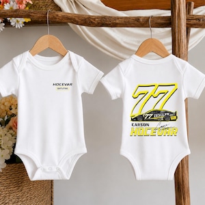 May include: Two white short-sleeve baby bodysuits hanging on wooden hangers. The left bodysuit features the text HOCEVAR. The right bodysuit displays the number 77, a race car graphic, the text CARSON HOCEVAR, and a signature in yellow and black.