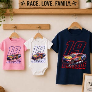 May include: A pink t-shirt, a white baby bodysuit, and a navy blue t-shirt, each featuring the number 19, a red race car graphic, and the text Chase Briscoe. The garments are displayed on wooden hangers against a light-colored wall.