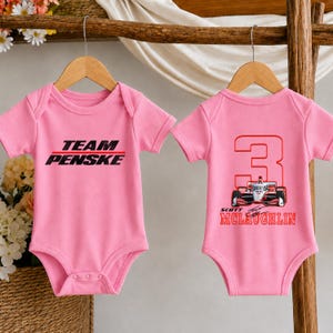 Puede incluir: Dos bodys de beb&eacute; rosas colgados en perchas de madera. El body izquierdo presenta el texto negro "TEAM PENSKE" con una l&iacute;nea roja. El body derecho muestra un n&uacute;mero "3" rojo grande, un gr&aacute;fico de coche de carreras y el texto rojo "SCOTT MCLAUGHLIN".