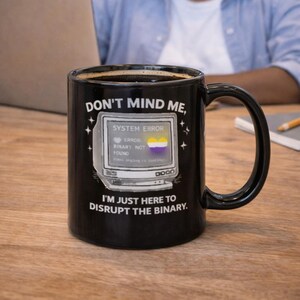 Può includere: Tazza in ceramica nera con la scritta "DON'T MIND ME, I'M JUST HERE TO DISRUPT THE BINARY." La tazza presenta una grafica di computer retrò con le parole "SYSTEM ERROR" e una bandiera non binaria. La tazza è su un tavolo di legno.