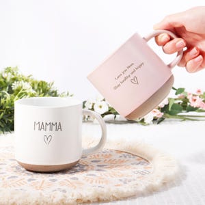 May include: Two ceramic mugs, one white and one pink, with brown bases. The white mug has the word "MAMMA" and a heart symbol. The pink mug has the text "Love you Mom, Stay healthy and happy." Both mugs have a handle.