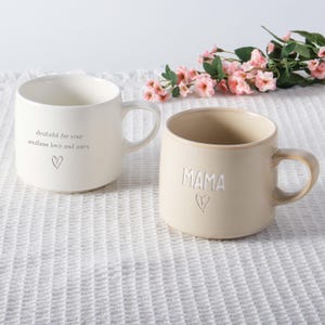 Puede incluir: Dos tazas de cerámica. Una es blanca con el texto "Grateful for your endless love and care" y un corazón. La otra es beige con la palabra "MAMA" y un corazón. Flores rosas en el fondo.