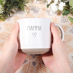 以下が含まれることがあります： 前面に「MAMMA」の文字とハートのシンボルが刻まれた白いセラミックマグ。マグカップの底は薄茶色です。マグカップには取っ手があり、両手で持っています。