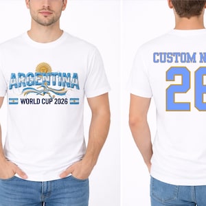 May include: White t-shirt featuring the word "ARGENTINA" in blue, with a sun graphic and the text "WORLD CUP 2026". The back of the shirt displays "CUSTOM NAME" and the number "26" in blue.