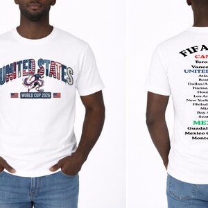 May include: White t-shirt with "UNITED STATES" in a patriotic design and "WORLD CUP 2026" text. The back of the shirt lists host cities in Canada, the United States, and Mexico for the FIFA World Cup 2026.