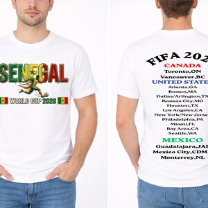 May include: White t-shirt featuring "SENEGAL WORLD CUP 2026" in green, red, and yellow, with a soccer player graphic. The back of the shirt lists FIFA 2026 host cities in Canada, the United States, and Mexico.