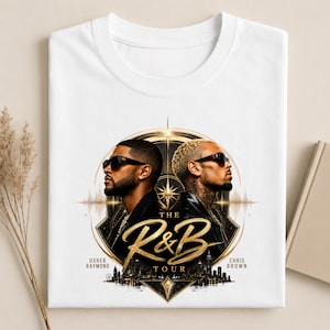 May include: A white t-shirt featuring a graphic print of Usher Raymond and Chris Brown wearing sunglasses. The design includes a gold circular frame, a city skyline silhouette, and the text "THE R&B TOUR" in a stylized gold script font.