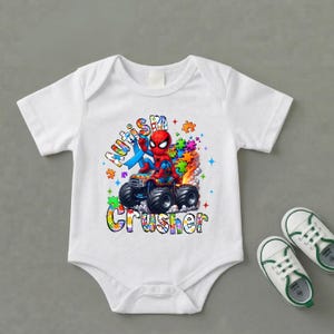 Puede incluir: Body de beb&eacute; blanco con un gr&aacute;fico colorido de Spider-Man conduciendo un cami&oacute;n monstruo. El texto "Autism Crusher" se muestra en una fuente colorida. Un par de zapatos de beb&eacute; blancos y verdes est&aacute;n al lado del body.