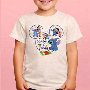 May include: A cream-colored short-sleeve t-shirt featuring a graphic of Stitch and Lilo inside a Mickey Mouse ear outline. The design includes the text "ohana means Family" in blue script, surrounded by small yellow stars and tropical flowers.