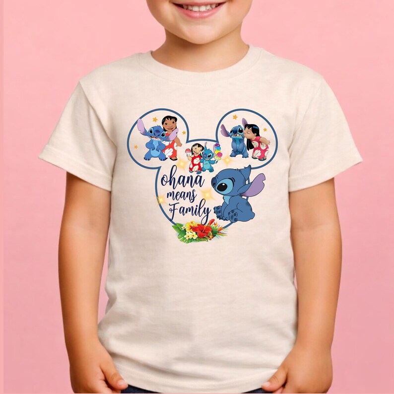 May include: A cream-colored short-sleeve t-shirt featuring a graphic of Stitch and Lilo inside a Mickey Mouse ear outline. The design includes the text "ohana means Family" in blue script, surrounded by small yellow stars and tropical flowers.