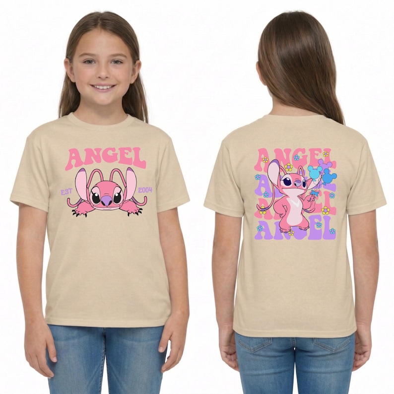 May include: A beige short-sleeved t-shirt featuring a pink Angel character graphic. The front displays the text ANGEL EST 2009. The back shows the character standing with flowers and mouse-ear shapes, with the word ANGEL repeated in pink and purple.