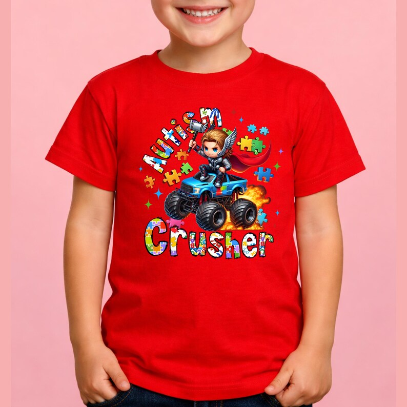 Puede incluir: Camiseta roja con un gr&aacute;fico colorido de un superh&eacute;roe conduciendo un cami&oacute;n monstruo azul. El texto de la camiseta dice "Autism Crusher" con acentos de piezas de rompecabezas. El dise&ntilde;o es vibrante y llamativo.