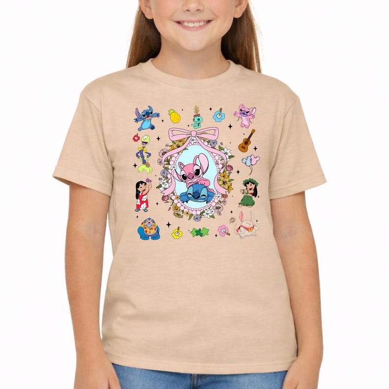 May include: A light tan short-sleeved t-shirt featuring a central oval frame decorated with flowers and a pink bow, containing blue and pink cartoon characters. Surrounding the frame are various small colorful cartoon icons and characters.