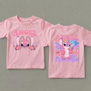 May include: A light pink short-sleeve t-shirt featuring a graphic of the character Angel. The front displays the text ANGEL EST 2004. The back shows Angel surrounded by small colourful flowers and blue mouse-ear shapes with the word ANGEL repeated.