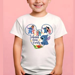 May include: A white short-sleeved t-shirt featuring a graphic of Stitch and Lilo inside a Mickey Mouse ear outline. The design includes the text "ohana means Family" in blue script, surrounded by small yellow stars and tropical hibiscus flowers.