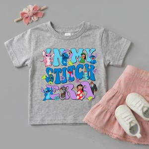 Pode incluir: Uma t-shirt cinzenta mesclada de manga curta com uma impress&atilde;o gr&aacute;fica colorida com o texto "IN MY STITCH ERA" rodeado por v&aacute;rias personagens de desenhos animados da franquia Lilo e Stitch, incluindo Stitch, Angel e Lilo, al&eacute;m de flores tropicais.