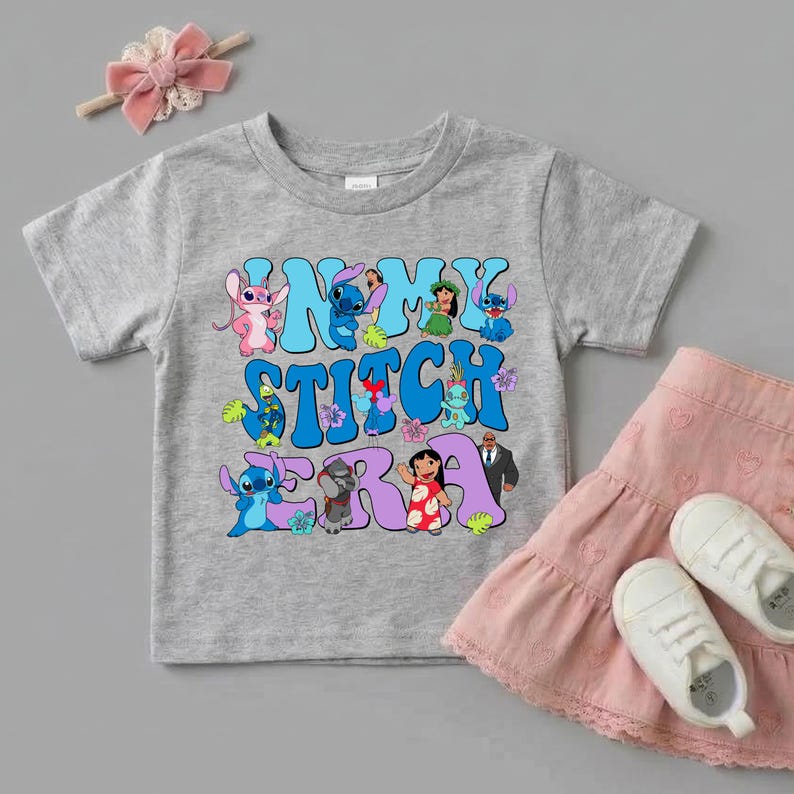 Pode incluir: Uma t-shirt cinzenta mesclada de manga curta com uma impress&atilde;o gr&aacute;fica colorida com o texto "IN MY STITCH ERA" rodeado por v&aacute;rias personagens de desenhos animados da franquia Lilo e Stitch, incluindo Stitch, Angel e Lilo, al&eacute;m de flores tropicais.