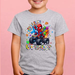 Puede incluir: Una camiseta gris con un gr&aacute;fico colorido de Spider-Man conduciendo un cami&oacute;n monstruo. El dise&ntilde;o incluye las palabras "Autism Crusher" y piezas de rompecabezas. La camiseta la lleva un ni&ntilde;o.