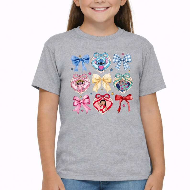 May include: A heather gray t-shirt featuring a central graphic print of various colorful bows and heart-shaped frames. The design includes illustrations of Stitch, Angel, and Lilo from Lilo and Stitch, interspersed with small stars and ribbons.